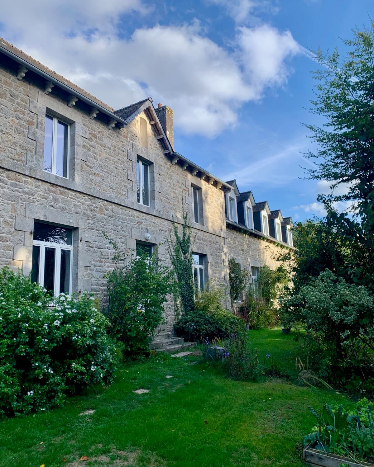 La façade en pierre de la Convergence des Loutres et son jardin, un soir d'été